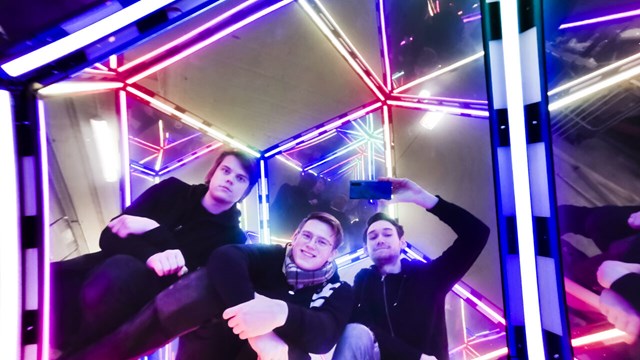 Tre unga män sitter inuti ett konstverk av speglar och neonljus
