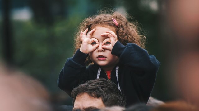 Ett litet barn sitter på en vuxens axlar och låtsas att fingrarna är glasögon